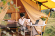© Ananass - Young woman works on laptop while sitting relaxed on chair by the campfire, traveling with tent. Concept of remote work and escape to nature