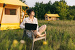 © Ananass - weekend work, Woman starts her morning working on a laptop while sitting near a tent in a glamping site for tourists