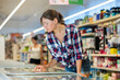 Interested young woman standing near refrigerated display cases choosing frozen food in supermarket..