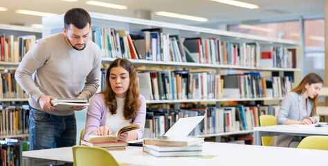 Canvas Print - Guy and a girl students studying at the university, preparing for the exam in the library studying on a laptop and looking ..for sources of information in books