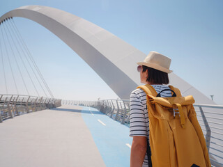 asian happy traveler woman with hat and yellow backpack enjoys stunning panoramic view of Dubai Creek Canal and famous tallest skyscraper Burj Khalifa, walk by Tolerance Bridge