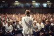 A woman in a white business suit is giving a speech on stage during a seminar. A lot of people in the blurred background. Shot from her back.