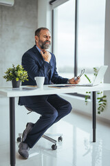 Wall Mural - Mature man having stressful time working on laptop.  Man working with laptop and paperwork with stress. Exhausted businessman using laptop at work and sitting by the desk while