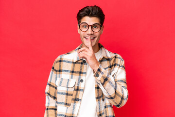 Wall Mural - Young caucasian handsome man isolated on red background showing a sign of silence gesture putting finger in mouth