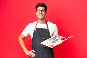 Wall Mural - Young artist caucasian man holding a palette isolated on red background posing with arms at hip and smiling