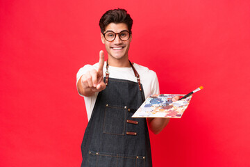 Wall Mural - Young artist caucasian man holding a palette isolated on red background showing and lifting a finger