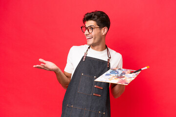 Wall Mural - Young artist caucasian man holding a palette isolated on red background with surprise expression while looking side