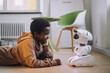 © Maskot - Smiling boy talking with AI robot while lying on floor in innovation lab