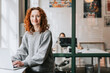© Maskot - Portrait of smiling redhead businesswoman with laptop at window sill in office