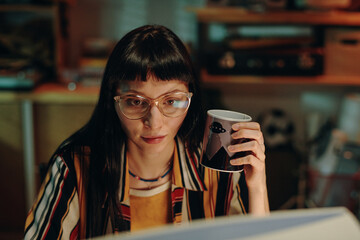 young woman in eyeglasses drinking coffee and working over new project on computer