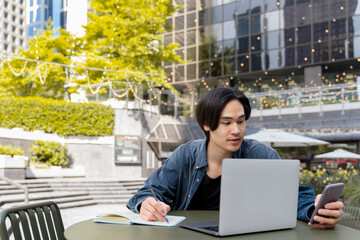 Wall Mural - Pensive asian male manager working outdoors, using smartphone, laptop, writing in notebook