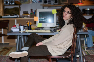 portrait of young programmer with curly hair looking at camera sitting at his workplace with compute