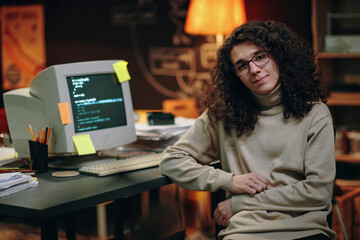 portrait of young developer with long curly hair looking at camera while sitting at his workplace wi