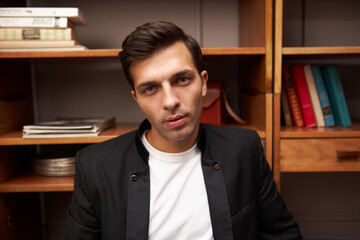 Closeup indoor portrait of handsome elegant caucasian male lawyer sitting against wooden bookshelf at his office, looking at camera, wearing black jacket and white t-shirt. Businessperson at work