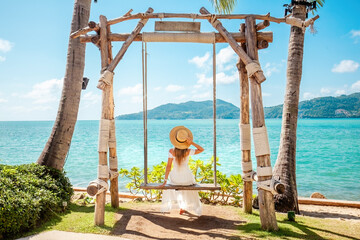 Relaxation and happiness of woman in a straw hat and white dress sitting on a swing with the turquoise sea on background. Tropical paradise, summer holidays and vacation.