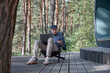 a young man with a beard and in a cap sits in nature against the backdrop of a pine forest and works in a laptop with a phone in his hands