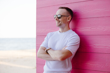 Wall Mural - Handsome smiling man posing with sunglasses leaning on pink wall, arms crossed