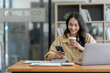 © crizzystudio - Asian businesswoman holding coffee cup and mobile phone sitting for break while working using laptop with paperwork, calculator making plan analyzing financial report business plan investment.