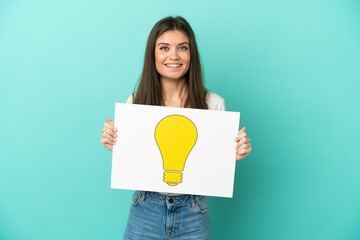 Wall Mural - Young caucasian woman isolated on blue background holding a placard with bulb icon with happy expression