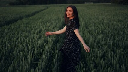 Wall Mural - A girl in a dress cheerfully runs through a field with ears of wheat. A young woman is running away turning around and smiling at the camera. A woman spends the summer in the village, farm