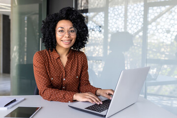 Portrait of mature adult successful businesswoman, african american woman at workplace smiling and looking at camera, confident boss working at workstation inside office, using laptop.