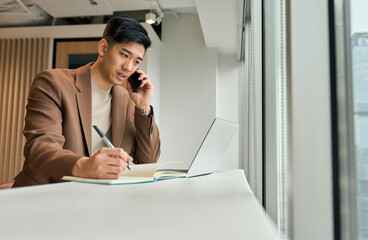 Busy Asian young business man sales manager making phone call using laptop in office. Japanese businessman talking on cellphone working consulting client discussing online digital financial data.