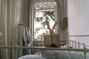 Woman reading a book in bed, lying on her stomach smiling happy and relaxed on a leisure day at home