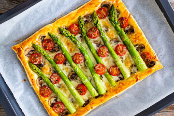 Wall Mural - Baked puff pastry with green asparagus with mozzarella cheese, mushrooms and cherry tomatoes on baking paper on wooden table
