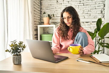 A young woman, focused and productive, working at her home desk with a laptop, sipping coffee, and feeling refreshed.