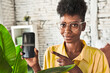 © Asier - African woman in her living room, showing a plant-related app advertisement on her phone.