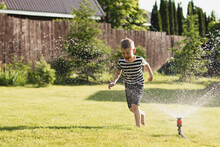 A Little Wet Boy Runs Barefoot On The Lawn Next To The Sprinkler. The Concept Of A Happy, Carefree Childhood And Vacation