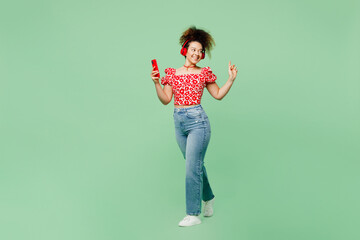 Wall Mural - Full body young woman she wearing casual clothes red blouse headphones listen music hold in hand use mobile cell phone isolated on plain pastel light green color background studio. Lifestyle concept.