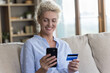 © fizkes - Positive satisfied senior woman holding banking credit card, smartphone, using modern Internet technology, commerce app, for shopping, payment from home, smiling, laughing, sitting on couch