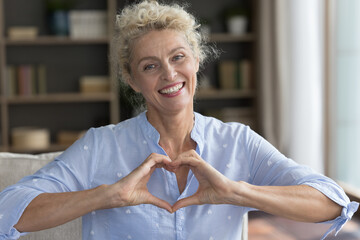 Cheerful positive pretty mature lady applying finger heart shaped hands to chest, looking at camera, smiling, posing at home, showing romantic gesture of love, happiness, peace, charity