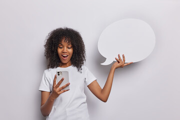 Studio shot of curly haired woman has surprised expression keeps mouth opened stares in smartphone holds blank speech bubble wears casual t shirt isolated over white background. Communication concept