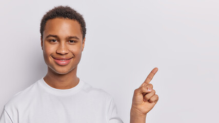Wall Mural - Cheerful dark skinned man promots product that demands discovery smiles pleasantly points index finger on copy space gives advice or advertises product dressed in casual t shirt isolated on white wall