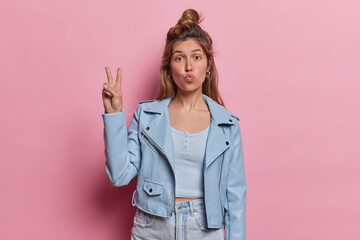 Wall Mural - Lovely dark haired woman with hair bun keeps lips folded makes peace gesture expresses positive emotions and joy dressed in blue jacket and jeans isolated over pink background. Body language concept