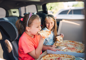 Portrait of two positive smiling sisters eating just cooked italian pizza sitting in child car seats on car back seat. Happy childhood, fastfood eating or auto jorney lunch break concept image.