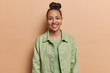 Portrait of cheerful Latin woman with hair bun smiles toothily stands carefree against brown background dressed in green shirt expresses positive emotions isolated. Happy feelings and emotions