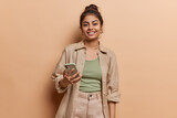 Fototapeta  - Studio shot of cheerful Indian girl with hair bun uses mobile phone for making video call or broadcasting livestream wears shirt and trousers isolated over brown background looks at device screen