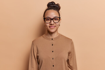 Canvas Print - Portrait of pretty schoolgirl pleased to have summer holidays at last wears transparant eyeglasses and jumper poses against brown background. Satisfied female student passed all exams successfully