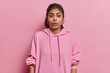 Stupefied dark haired Indian woman stands speechless has shocked facial expression looks at something unbelievable dressed in casual hoodie isolated over pink background. Human reactions concept