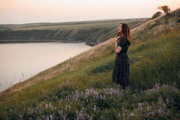 Wall Mural - Portrait of a beautiful young brunette woman in a summer dress at sunset among beautiful nature. The girl fixes her hair and looks into the distance. Place for text.