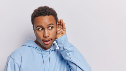 Wall Mural - Surprised dark skinned man tries to overhear someone holds hand near ear tries to understand words eavesdropping has stunned expression wears blue sweatshirt isolated over white background copy space