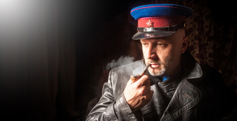 Officer from army of USSR. Man in NKVD cap. Investigator smokes pipe. KGB officer in leather coat. Symbol of USSR on cap. Member of soviet army. Portrait of man officer of KGB of USSR