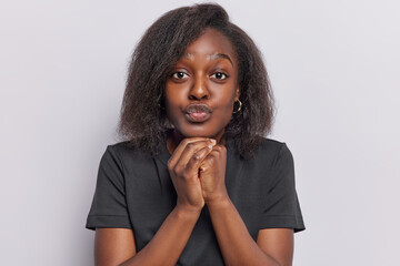 Wall Mural - Studio shot of dark skinned ethnic woman keeps hands under chin has lips folded looks with romantic expression at camera wears casual black t shirt isolated over white background adores something