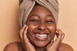 © wayhome.studio  - Close up portrait of dark skinned woman smiles toothily touches cheeks gently has soft clean skin after showering wears wrapped towel on head isolated over brown background takes care of herself