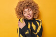 Lovely happy curly haired woman embraces herself daydreamsand embraces herself with both hands recalls romantic event wears comfortable jumper isolated over yellow background. Egoism concept