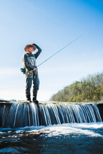 A Young Woman In Wader Fly Fishing Near A Small Waterfall On The French Broad River Near Asheville, North Carolina