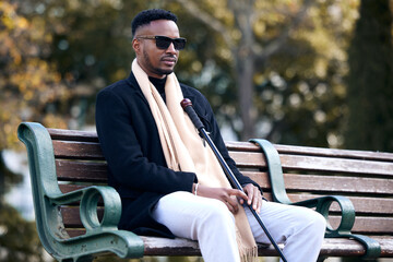 Blind black man, park and sitting with sunglasses, walking stick and relax by trees, peace and thinking. African guy, vintage fashion and young person with disability, glasses and travel in nature